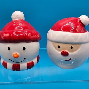 May include: A set of two ceramic Christmas salt and pepper shakers. One shaker is a snowman with a red hat and scarf. The other shaker is Santa Claus with a red hat and white beard.