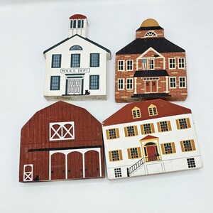 May include: Set of four wooden miniature buildings painted in various colors. The buildings include a red barn, a white house with a red door, a white police station with a blue door, and a white schoolhouse with a red door. Each building has a black cat painted on the front.