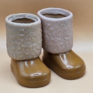 May include: Two brown ceramic boots with white fuzzy trim. The boots are shiny and have a rounded toe.