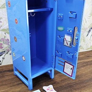 May include: A blue miniature locker with an open door, featuring a shelf, a hanging bar, and various stickers. The door has a small notepad with the text "Practice Tuesday Night". Several small decorative frames are in front of the locker.