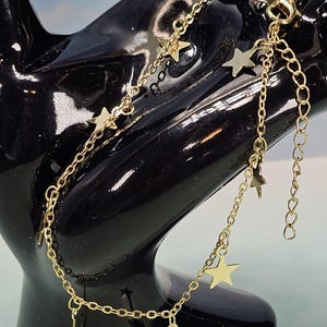 May include: Gold-colored anklet with multiple strands and dangling star charms. The anklet is displayed on a black surface. The stars are evenly spaced along the delicate chain, creating a celestial design.