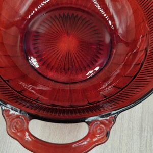 May include: A red glass bowl with two handles. The bowl has a textured, sunburst design on the inside and a black rim.