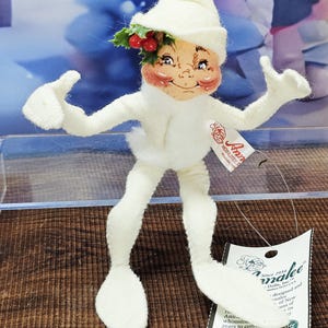 May include: A white Annalee doll with a whimsical design. The doll has a pointed hat, rosy cheeks, and a sprig of holly with red berries. The doll is wearing white gloves and shoes. The tag reads "Annalee Dolls, Inc."