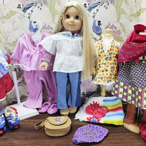 May include: A doll with long blonde hair stands among various outfits and accessories. Clothing includes a white blouse, blue jeans, a pink pajama set, and a floral dress. Accessories include roller skates, a purse, and a crocheted hat.