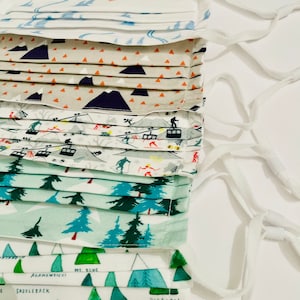 May include: Five fabric face masks with different winter-themed prints. The masks are white with various patterns including mountains, trees, and ski lifts. The bottom mask has the names of mountains printed on it: Saddleback, Mt. Blue, and Bigelow.