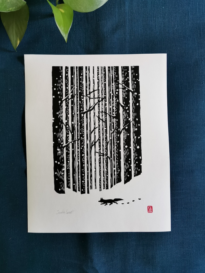 May include: Black and white linocut print depicting a snowy forest scene. A fox walks through the snow, leaving tracks. The artwork features vertical tree trunks, falling snow, and bare branches. Signed by the artist, Santa Lucat, with a red stamp.