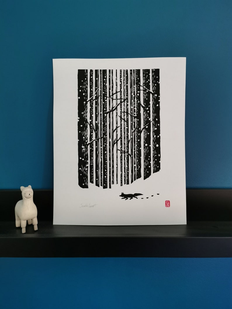 May include: Black and white art print depicting a winter forest scene with falling snow and a fox. The print features vertical tree trunks, a fox silhouette, and the artist's signature. A small white llama figurine is on the left.