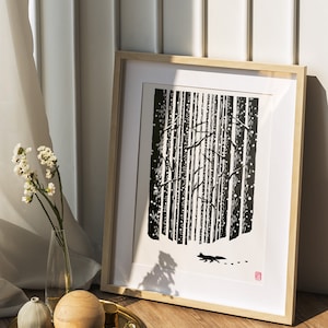 May include: Framed black and white art print depicting a forest scene with a fox silhouette. The artwork is in a light wood frame, leaning against a white wall. A decorative tray with wooden spheres and a vase of flowers sits in front.