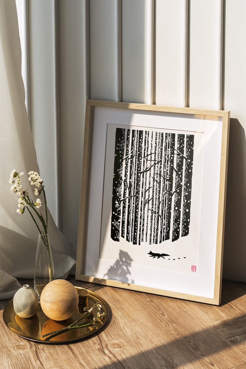 May include: Framed black and white art print depicting a forest scene with a fox silhouette. The artwork is in a light wood frame, leaning against a white wall. A decorative tray with wooden spheres and a vase of flowers sits in front.