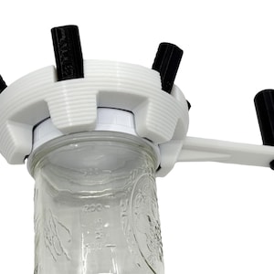 May include: A white jar opener with black handles and a crank. The opener is designed to fit over the lid of a glass jar. The jar is clear with measurements in ounces.