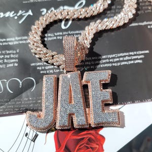 Puede incluir: Un collar de cadena de oro rosa con un colgante que deletrea "JAE" en letras grandes y en bloque. Las letras y la cadena están incrustadas con gemas claras y brillantes. El collar se exhibe sobre una superficie reflectante.
