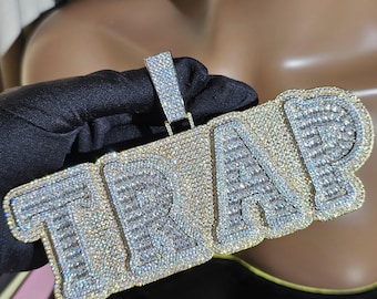 May include: A gold pendant with the word "TRAP" spelled out in large, bold letters. The letters are covered in clear, sparkling rhinestones.