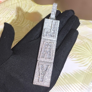 May include: A rectangular pendant with the word "FOREVER" spelled out in sparkling white gemstones. The pendant is made of silver-colored metal and is held by a black gloved hand.