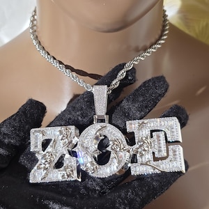 May include: A silver-toned pendant necklace featuring the letters ZOE in a block font. The letters are encrusted with numerous small, clear, rectangular-cut stones and decorated with raised, metallic rose floral motifs across the surface.