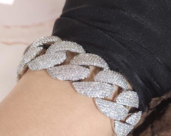 May include: A silver-colored, chunky link bracelet. The bracelet is covered in small, sparkling stones, creating a dazzling effect. The bracelet is worn on a wrist, with a black sleeve in the background. The bracelet's design is bold and eye-catching.