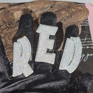 May include: Three silver rings spelling out the word "RED". Each letter is encrusted with small, sparkling stones. The rings are displayed on a black surface, with a wooden background.