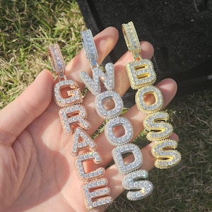 May include: Three pendant necklaces spelling out "GRACE", "WOODS", and "BOSS". The pendants are made of gold, silver, and rose gold-toned metal, each encrusted with clear, sparkling gemstones. The necklaces are held against a backdrop of green grass.