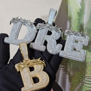 May include: Three gold and silver pendants with a crown of thorns design. Each pendant is a letter, spelling out the word "DRE". The pendants are covered in sparkling stones.