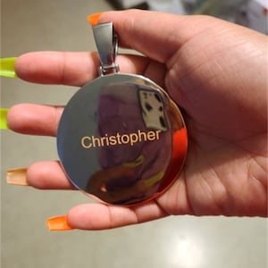May include: A silver round pendant with a polished finish and the name "Christopher" engraved in gold lettering.