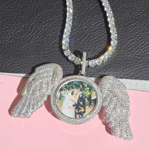 Può includere: Una collana con pendente tempestata di diamanti con una cornice rotonda e due ali ricoperte di diamanti. La collana ha una catena tennis con piccole pietre trasparenti. Il pendente mostra una foto di una coppia che si bacia. Lo sfondo è nero e rosa.
