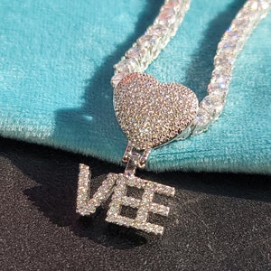May include: A silver-toned necklace with a heart-shaped pendant and the letters "VEE". The necklace chain and pendant are encrusted with small, clear gemstones. The heart pendant is attached to the chain with a small clasp.
