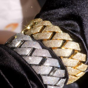 Peut inclure: Gros plan sur deux bracelets à maillons cubains incrustés de diamants. L'un est argenté, l'autre doré. Les bracelets sont présentés sur un fond en tissu noir, avec la lumière se reflétant sur les pierres précieuses.