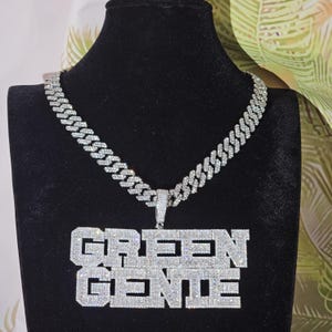 Puede incluir: Un collar de cadena de plata con un gran colgante que dice "GREEN GENIE" en letras mayúsculas. El colgante y la cadena están cubiertos de piedras de circonita cúbica transparentes.