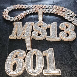May include: A gold chain necklace with two pendants. The first pendant is a large, diamond-encrusted pendant with the letters "MS13". The second pendant is a large, diamond-encrusted pendant with the numbers "601".