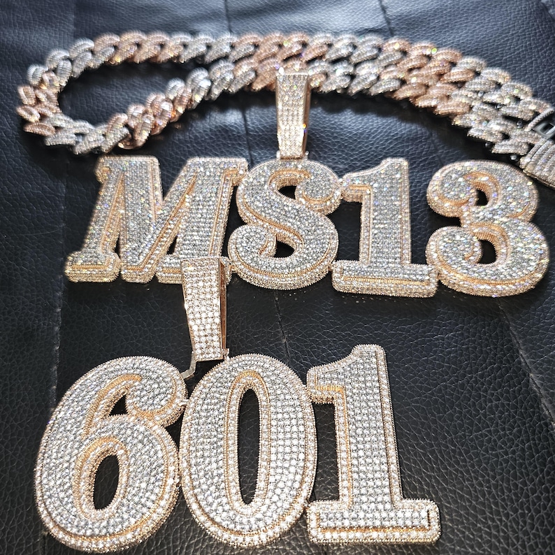May include: A gold chain necklace with two pendants. The first pendant is a large, diamond-encrusted pendant with the letters "MS13". The second pendant is a large, diamond-encrusted pendant with the numbers "601".