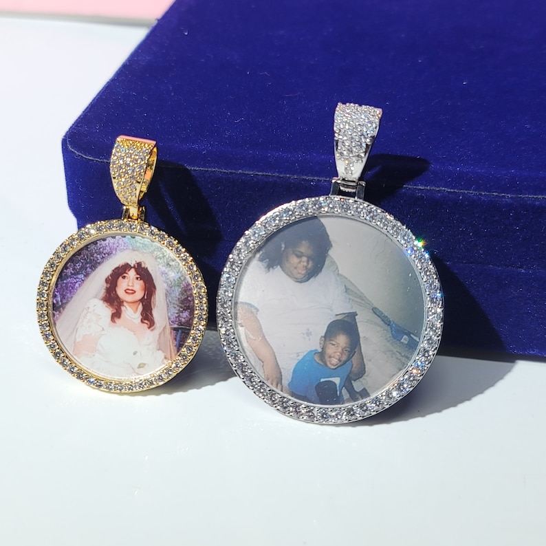 May include: Two silver and gold pendants with a round photo frame encrusted with clear cubic zirconia stones. The pendants are on a blue velvet background.