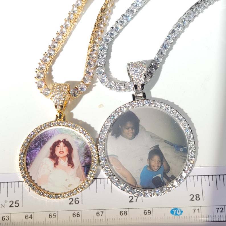May include: Two silver and gold chains with round photo pendants. The pendants are encrusted with clear stones and feature photos of people. The chains are laid out on a ruler with markings from 25 to 72.