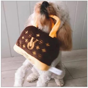 May include: A white dog with brown fur playing with a brown and yellow plush toy shaped like a handbag. The handbag has a brown and gold pattern with a large "V" in the center.