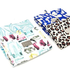 Puede incluir: Cuatro bolsas de tela reutilizables para la compra con fondo blanco y diferentes estampados. Las bolsas están dobladas y tienen una etiqueta de metal plateado con la palabra "clutch" en ellas.
