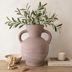 May include: A large, textured, mauve-colored vase with two handles, filled with green leafy branches. A small, off-white bowl with wooden beads and a tassel, a candle, and a book with the text "LIVE BEAUTIFULLY" are on a wooden surface.