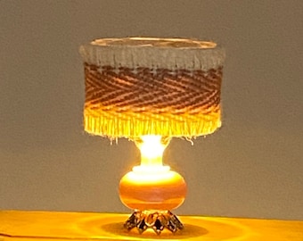 Op de afbeelding: Een miniatuur tafellamp met een bruine en witte geweven lampenkap en een roze voet. De lamp is aan en verlicht de tafel.