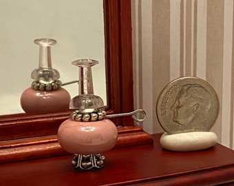 May include: A miniature pink glass lamp with a clear glass chimney and a silver base. The lamp is sitting on a wooden dresser with two drawers and gold handles. A silver dime is sitting on a white stone next to the dresser.