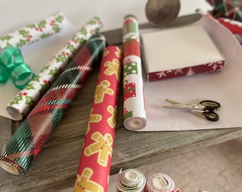 May include: Assortment of Christmas wrapping paper rolls with festive designs like holly, tartan, gingerbread men, and gift motifs. A small box with a Christmas tree pattern and scissors are also visible, ideal for gift wrapping.