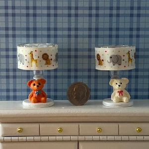 Può includere: Due lampade da tavolo bianche con una base a forma di orsacchiotto bianco e un paralume bianco con stampa di animali colorati. Le lampade sono su una cassettiera bianca con pomelli dorati.