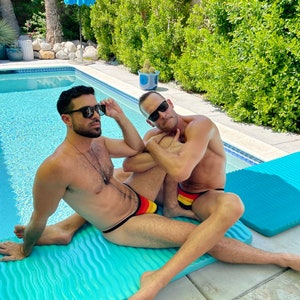 May include: Two people wearing sunglasses and swim briefs with black straps and a yellow, red, and black design, lounging on a turquoise pool float. The background includes a swimming pool, lush greenery, and a blue umbrella.