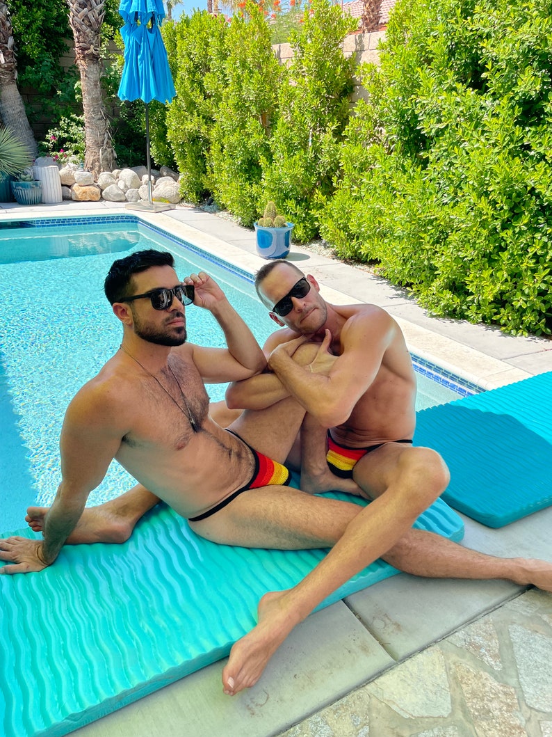May include: Two people wearing sunglasses and swim briefs with black straps and a yellow, red, and black design, lounging on a turquoise pool float. The background includes a swimming pool, lush greenery, and a blue umbrella.