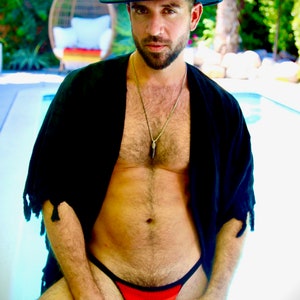 May include: A black fedora hat and a black open-front top are worn. A crochet thong in the colors of the German flag is visible. A silver necklace hangs around the neck. The background includes a pool and greenery.