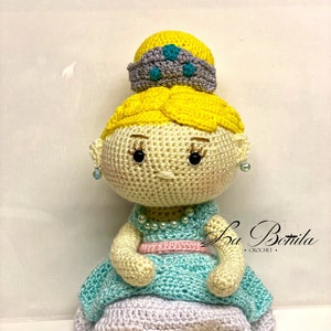 May include: A crocheted doll with a yellow bun, a blue crown, a light blue dress, and gray shoes. The doll is sitting on a white surface. The text "La Bonita Crochet" is visible in the background.