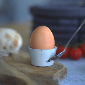 Könnte beinhalten: Ein grauer Eierbecher aus Beton mit einem braunen Ei darin. Ein silberner Löffel liegt am Rand des Eierbechers. Der Eierbecher steht auf einem Holzbrett.