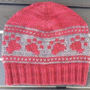 Puede incluir: Gorro de punto rojo con un diseño estampado en gris que presenta un motivo repetido de renos estilizados. El gorro tiene un puño de canalé.