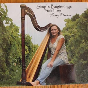 May include: CD cover featuring a woman with a harp. The harp is brown and gold, with a curved top. The woman is wearing a patterned top and jeans. The text on the cover reads "Simple Beginnings Solo Harp Nancy Eischen."