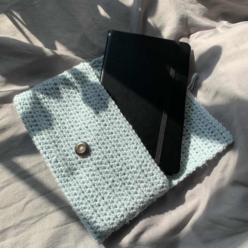 May include: A light blue crocheted clutch with a black notebook inside. The clutch has a single button closure.