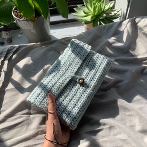 May include: A light gray crocheted clutch with a flap closure and a single button. The clutch is lying on a gray fabric surface.