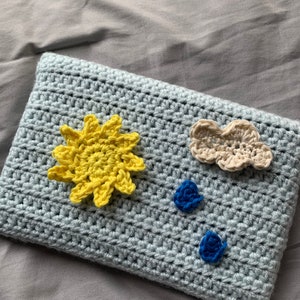 May include: A light blue crocheted pillow with a yellow sun, a beige cloud, and two blue flower embellishments.