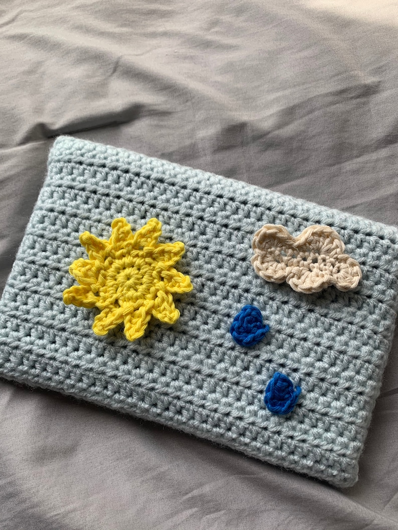 May include: A light blue crocheted pillow with a yellow sun, a beige cloud, and two blue flower embellishments.