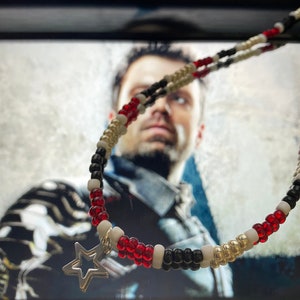 Peut inclure: Un collier de perles rouge, blanc et noir avec un pendentif en forme d'étoile argentée. Le collier est net, tandis qu'un visage de personne est flou en arrière-plan.